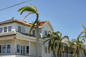 White coastal house with damaged roof and tall palm trees against a clear blue sky